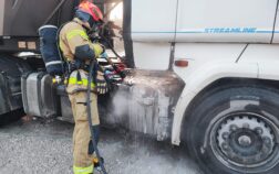 Widać kawałek ciągnika i naczepy auta ciężarowego, z dołu auta unosi się lekki dym. Przed autem stoi strażak w aparacie powietrznym i podaje wodę.