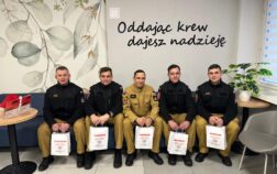 5 strażaków siedzi na kanapie w mundurach, w rękach trzymają torby z centrum krwiodawstwa. Nad ich głową na ścianie jest napis oddając krew dajesz nadzieję.