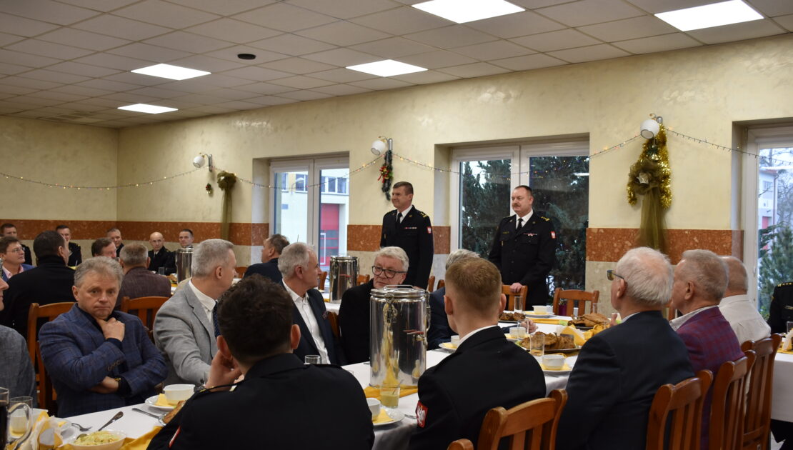 Strażacy w mundurach galowych i osoby cywilne siedzą przy stołach, jeden ze strażaków przemawia