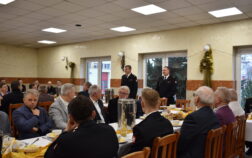 Strażacy w mundurach galowych i osoby cywilne siedzą przy stołach, jeden ze strażaków przemawia