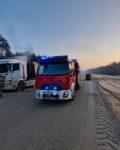3 Na drodze po lewej stronie stoi auto ciężarowe z nad którego unosi się lekki dym. obok po prawej stronie stoi samochód strażacki. Przed autami widać dwóch strażaków.