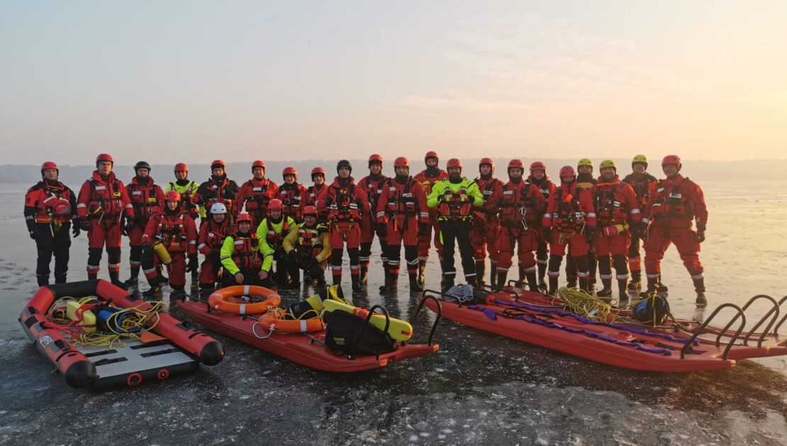 26 strażaków w specjalnych ubraniach stoją na lodzie, przed nimi leżą sanie i trapy do ratownictwa.