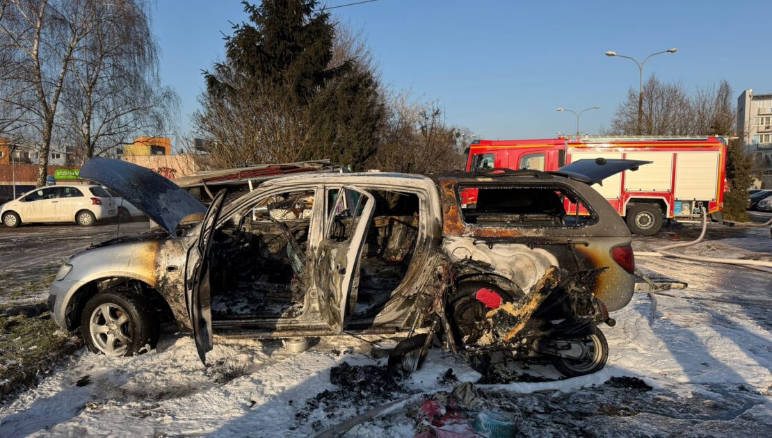 na parkingu stoi spalone auto osobowe pokryte pianą gaśniczą. W tle widać samochód strażacki