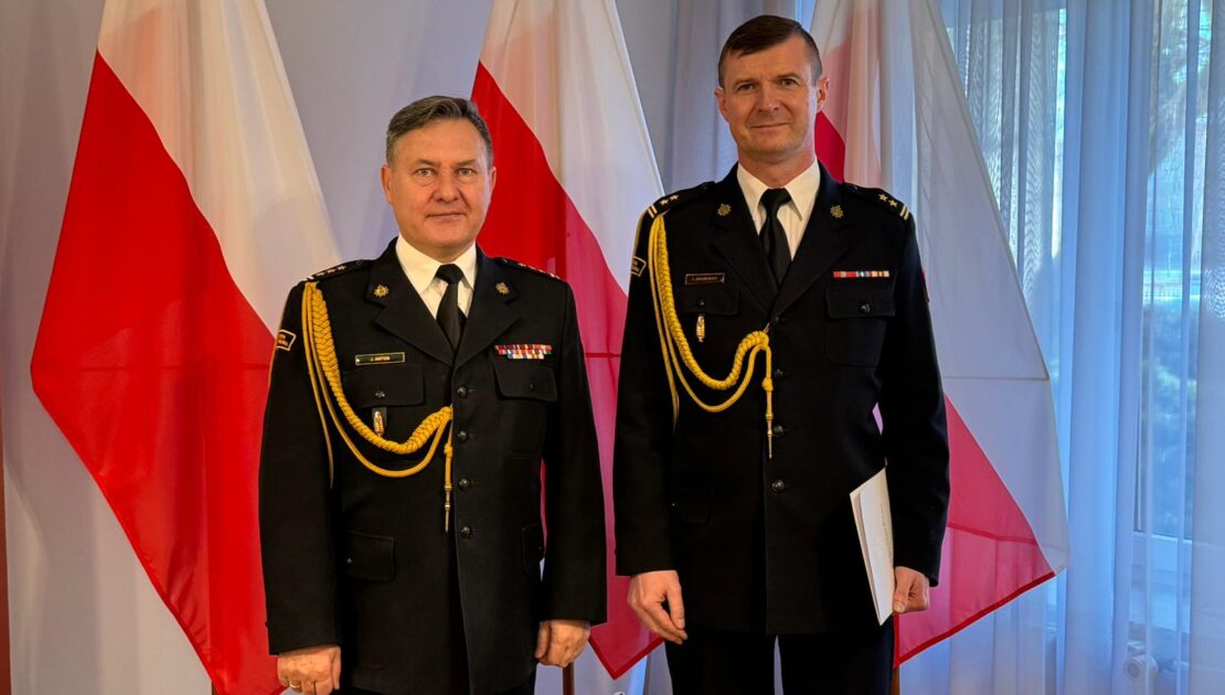 Dwóch strażaków w mundurach wyjściowych stoi obok siebie jeden z nich trzyma w ręku teczkę. W tle flaga Polski