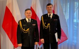 Dwóch strażaków w mundurach wyjściowych stoi obok siebie jeden z nich trzyma w ręku teczkę. W tle flaga Polski