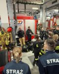 WhatsApp Image 2026-02-05 at 07.32.55 (1) W garażu stoją auta strażackie pomiędzy nimi stoją grupa młodzieżowej drużyny pożarniczej, jeden ze strażaków prezentuje sprzęt