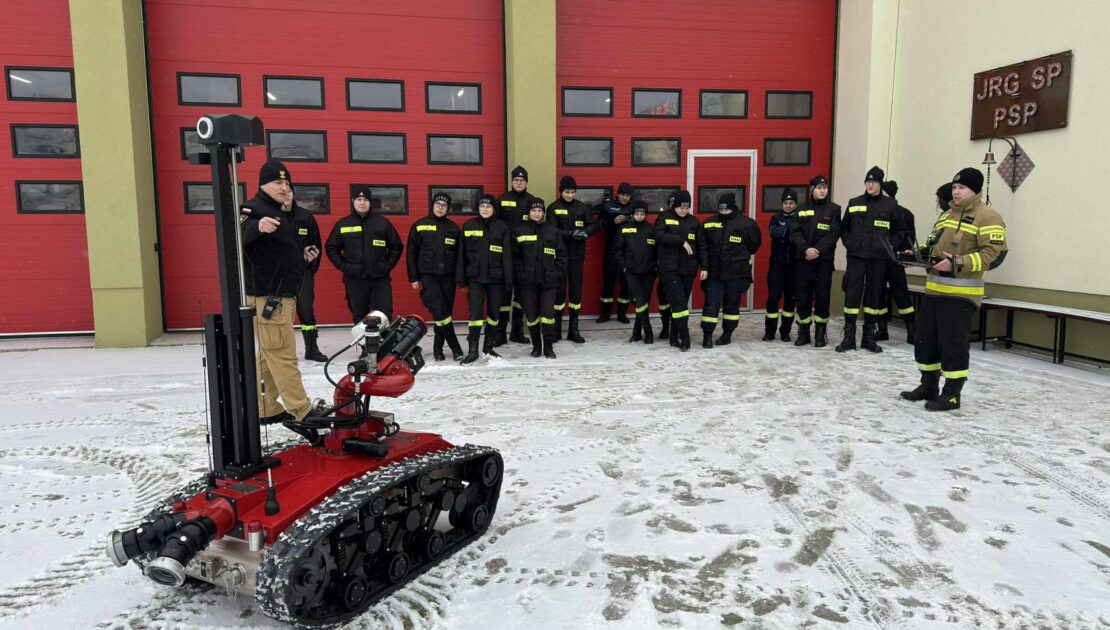 Grupa strażaków stoi przed bramami garażowymi, przed grupą stoi robot gaśniczy