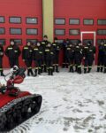 WhatsApp Image 2026-02-05 at 07.32.56 (5) Grupa strażaków stoi przed bramami garażowymi, przed grupą stoi robot gaśniczy