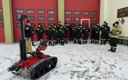 Grupa strażaków stoi przed bramami garażowymi, przed grupą stoi robot gaśniczy