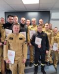 15 strażaków w mundurach koszarowych i służbowych stoją , część z nich trzyma teczki