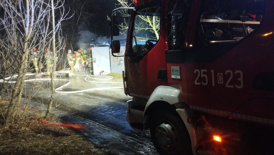 Po prawej stornie kabina pojazdu strażackiego przed nią w tle garaż znad którego unosi się dym