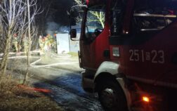 Po prawej stornie kabina pojazdu strażackiego przed nią w tle garaż znad którego unosi się dym