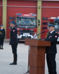 Komendant Szkoły stoi przy mównicy, w strażacy stojący przy maszcie, w tle samochody pożarnicze