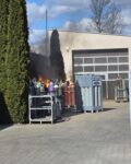 4 Na placu w koszu stoją butle, z których ulatnia się gaz i spala niewielkim płomieniem. W tle budynek garażowy