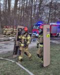 5 Dwóch strażaków w pełnym umundurowaniu specjalnym niesie tarcze balistyczne, w tle inni strażacy i samochód strażacki