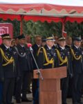 Goście honorowi stoją na trybunie pod namiotem, widać strażaków w mundurach wyjściowych i inne osoby
