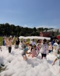 Grupa radosnych dzieci bawi się w ogromnej ilości białej piany na trawiastym placu. Po prawej stronie widoczna jest czarna armatka, która wyrzuca w górę gęsty strumień piany, tworząc nad dziećmi biały łuk pod bezchmurnym, błękitnym niebem