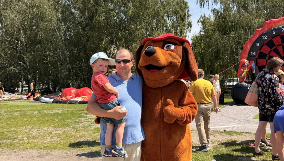 Maskotka dużego brązowego psa w czerwonej czapce kuca na trawie i przybija „piątkę” z małym chłopcem. W tle widoczne są duże, czerwono-czarne dmuchańce oraz ściana zielonych drzew