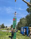 Dziecko w kasku i uprzęży stoi na szczycie bardzo wysokiej wieży zbudowanej z kilkunastu zielonych skrzynek. U dołu wieży, na drewnianej palecie, stoi instruktor w ciemnych spodniach i piaskowej koszulce, który obiema rękami asekuruje i stabilizuje konstrukcję . Bezpieczeństwo dziecka zapewnia lina asekuracyjna zwisająca z ramienia dźwigu, którego fragment widoczny jest u góry kadru na tle czystego, niebieskiego nieba . Zabawa odbywa się na trawiastym terenie w słoneczny dzień