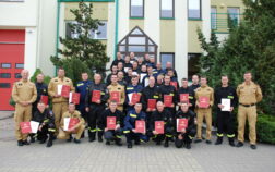 38 strażaków w mundurach koszarowych i służbowych stoją , część z nich trzyma teczki
