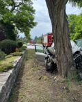 1 Rozbity srebrny samochód osobowy ze zmiażdżonym przodem po uderzeniu w grube drzewo na trawiastym poboczu. W tle, na drodze za pojazdem, widoczne są dwa wozy strażackie oraz radiowóz policyjny na sygnale.