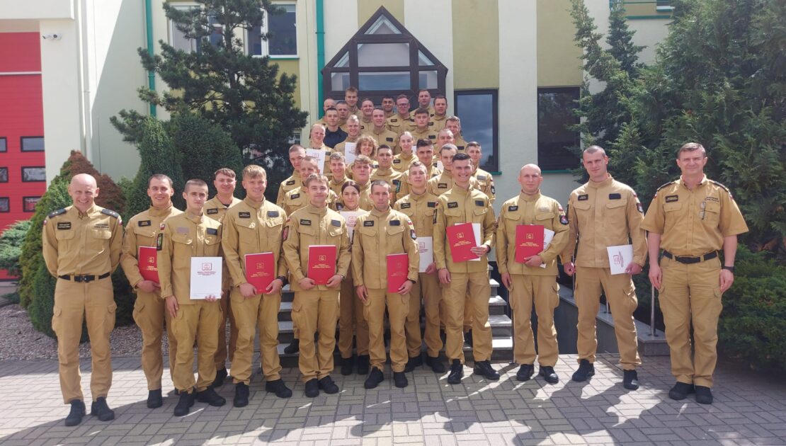 Duża grupa strażaków pozuje do zdjęcia grupowego przed budynkiem straży pożarnej w słoneczny dzień. Wielu z nich trzyma dokumenty lub certyfikaty, z których niektóre mają czerwone okładki. Strażacy są ubrani w mundury