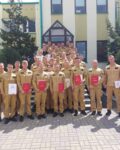 3 Duża grupa strażaków pozuje do zdjęcia grupowego przed budynkiem straży pożarnej w słoneczny dzień. Wielu z nich trzyma dokumenty lub certyfikaty, z których niektóre mają czerwone okładki. Strażacy są ubrani w mundury