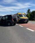 8 Czarny samochód terenowy z mocno uszkodzonym bokiem, otwartymi drzwiami i podniesioną maską stoi na jezdni po kolizji. Tuż obok pojazdu zaparkowana jest żółta karetka pogotowia ratunkowego na sygnale świetlnym. Akcja odbywa się na dwupasmowej drodze w słoneczny dzień.