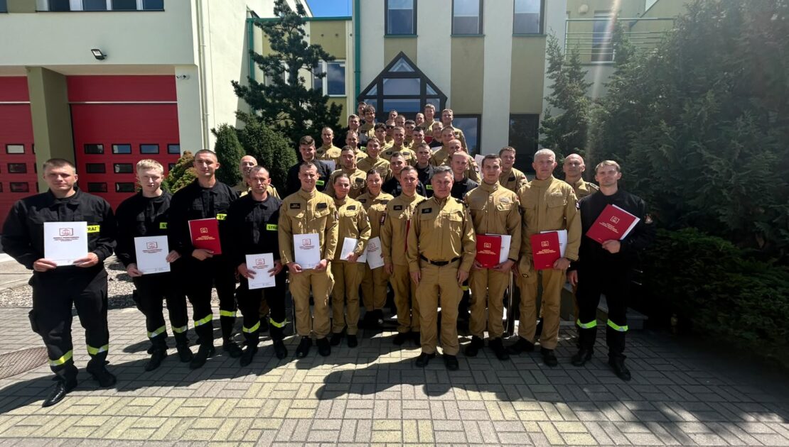 Duża grupa strażaków pozuje do zdjęcia grupowego przed budynkiem straży pożarnej w słoneczny dzień. Wielu z nich trzyma dokumenty lub certyfikaty, z których niektóre mają czerwone okładki. Strażacy są ubrani w mundury