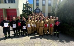 Duża grupa strażaków pozuje do zdjęcia grupowego przed budynkiem straży pożarnej w słoneczny dzień. Wielu z nich trzyma dokumenty lub certyfikaty, z których niektóre mają czerwone okładki. Strażacy są ubrani w mundury