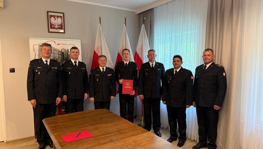 Uroczystość ślubowania w Państwowej Straży Pożarnej. W oficjalnym gabinecie stoi siedmiu strażaków w ciemnogranatowych mundurach galowych. Nowo przyjęty strażak w centrum trzyma czerwoną teczkę z aktem ślubowania. W tle widoczne są polskie flagi, godło na ścianie oraz duże okna z firanami, a na pierwszym planie drewniany stół konferencyjny.