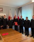WhatsApp Image 2026-07-29 at 13.01.18 Uroczystość ślubowania w Państwowej Straży Pożarnej. W oficjalnym gabinecie stoi siedmiu strażaków w ciemnogranatowych mundurach galowych. Nowo przyjęty strażak w centrum trzyma czerwoną teczkę z aktem ślubowania. W tle widoczne są polskie flagi, godło na ścianie oraz duże okna z firanami, a na pierwszym planie drewniany stół konferencyjny.